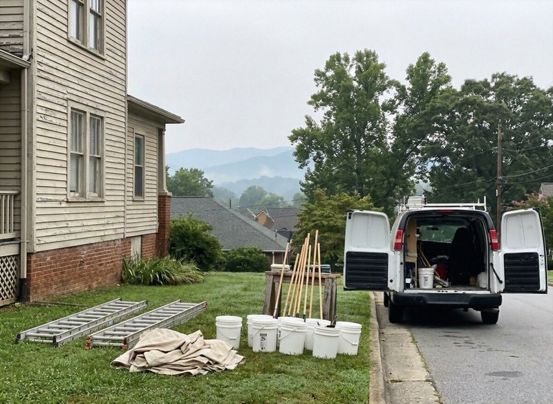 Ladders, drop cloths and paint buckets laid out on a lawn beside a white work van on the first morning of an exterior repaint